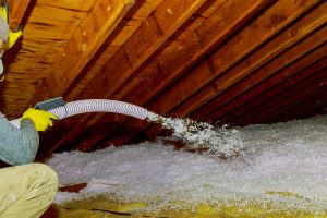 Technician spraying blown fiberglass insulation between trusses in attic.