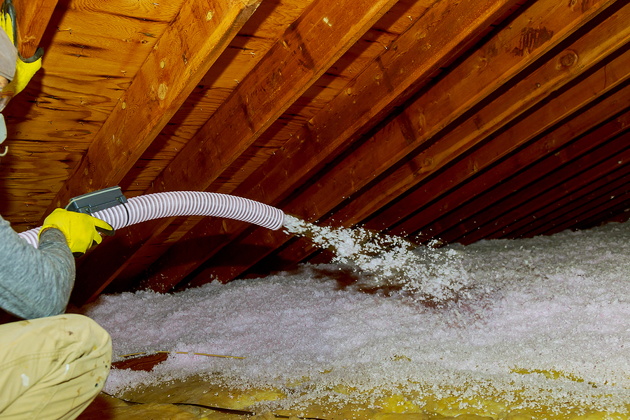 Technician spraying blown fiberglass insulation between trusses in attic.
